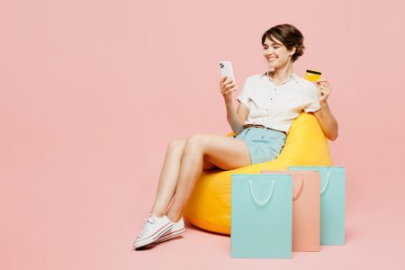 Full body happy young woman wear casual clothes hold bags sit in bag chair use mobile cell phone credit bank card shopping online isolated on plain pink background. Black Friday sale buy day concept