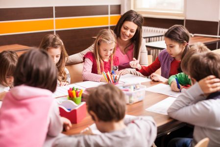 Children boys and girls sitting together around the table in classroom and drawing. With them is their young and beautiful teacher.